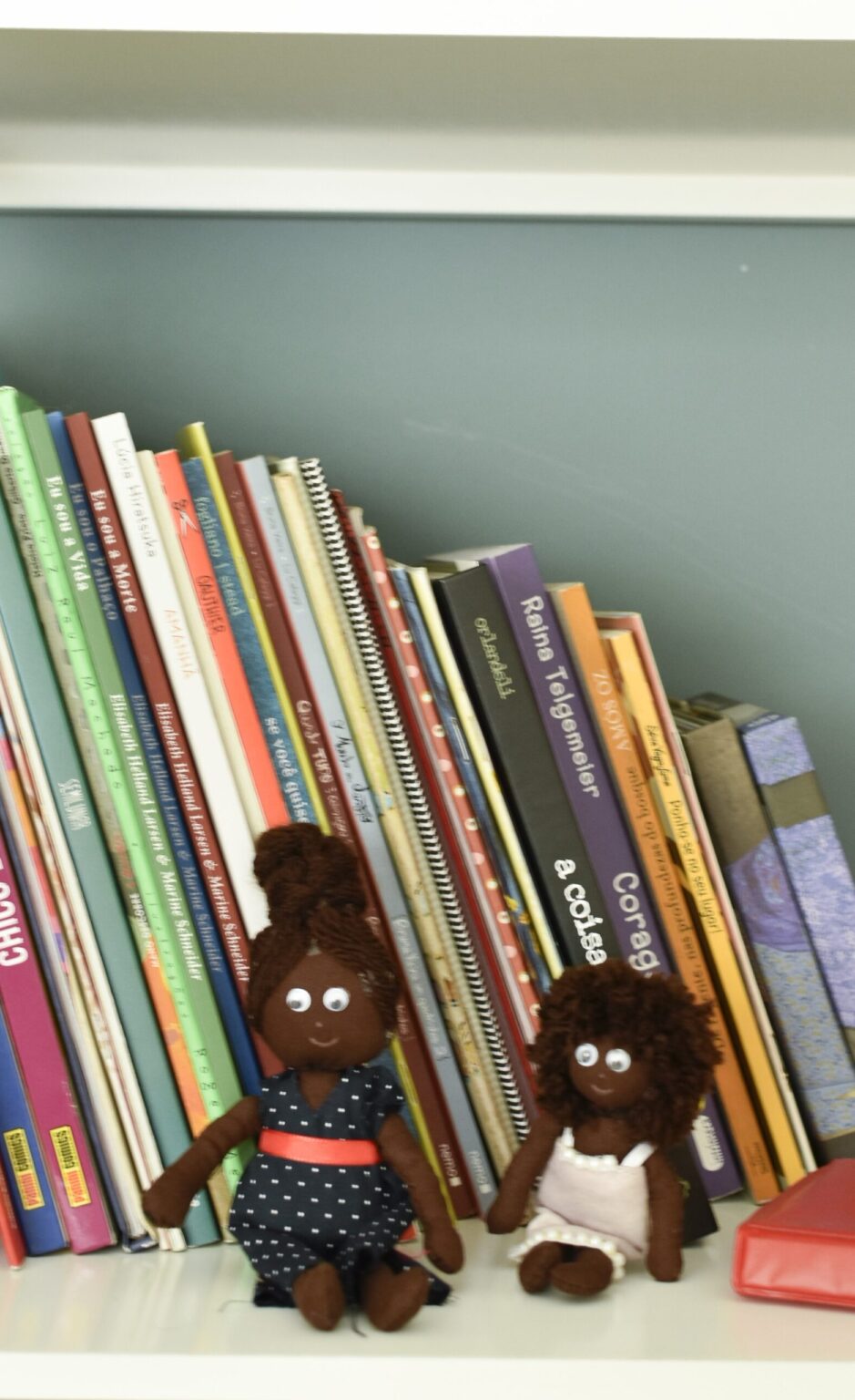 Bonecas negras em estante de livros infantojuvenis e jogos do consultório da psicóloga.